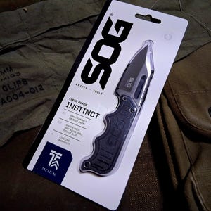 May include: A black SOG Instinct fixed blade knife in its packaging. The packaging is white with black text, including "SOG" and "INSTINCT". The knife has a black handle and a silver blade. The packaging also mentions a lifetime warranty.
