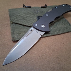 May include: A white ceramic mug with a black handle and the text "But First, Coffee" in a black, bold font. The mug is sitting on a wooden surface. The mug is approximately 4 inches tall.