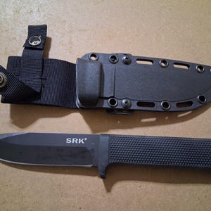 May include: A black SRK knife with a textured handle and a black sheath. The knife has a pointed blade and the text "SRK" is printed on the blade. The sheath has a belt loop and is secured with screws.
