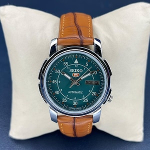 Puede incluir: Un reloj automático Seiko con esfera verde y caja plateada, con correa de cuero marrón. La esfera muestra el día y la fecha, con números y agujas blancos. El reloj se exhibe sobre un cojín blanco.