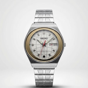 Può includere: Un orologio da polso automatico Seiko argentato con una lunetta dorata. Il quadrante bianco presenta un motivo a griglia, una lancetta dei secondi rossa e un'indicazione della data. Il cinturino è argentato.