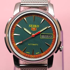 Peut inclure: Une montre automatique Seiko 5 vintage avec un cadran vert et un bracelet en maille argentée. Le cadran de la montre présente des accents rouges et orange, des aiguilles lumineuses et un affichage jour-date. Une montre classique.