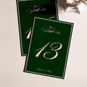 May include: Two dark green table number cards with white borders. The cards feature the words "Table No." in a script font and the numbers "1" and "13" in a soft pink color. The bottom of the card reads "Amanda & Jason".