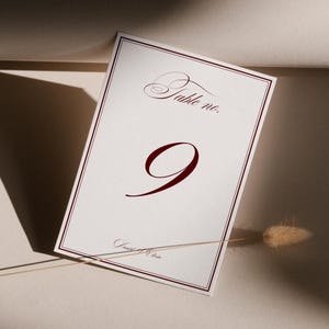 Puede incluir: Una tarjeta blanca con el número de mesa con un borde burdeos y el número 9 en una fuente elegante y en negrita. Las palabras "Table no." están en la parte superior, y un nombre en la parte inferior. Una flor seca está en primer plano.