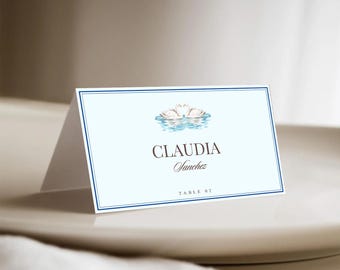 May include: A light blue place card with a dark blue border. The card features a watercolor illustration of two swans on water and the names "Claudia Sanchez" in elegant script. The text "TABLE 02" is also present.