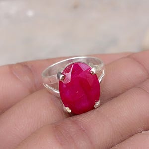 May include: A silver ring featuring a large, oval-shaped, faceted deep pink gemstone set in a four-prong mounting. The band of the ring is split into two thin silver segments that connect to the central setting, showcasing a polished metallic finish.