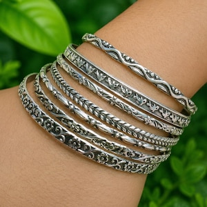May include: A set of six silver-toned metal bangles worn together. Each bracelet features a distinct embossed pattern, including floral motifs, braided textures, and stylized bird designs, all finished with an oxidized look to highlight the details.
