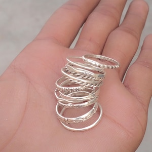 Op de afbeelding: Een stapel zilveren ringen met diverse ontwerpen, waaronder gedraaide, kralen en patronen. De ringen worden op een hand getoond, waarbij hun verschillende texturen en stijlen worden benadrukt. De ringen zijn een mix van dunne en iets dikkere banden.