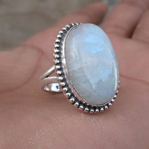 May include: Oval moonstone ring with a silver band. The moonstone has a pearly white color with blue iridescence. The ring's bezel is detailed with small silver beads. A split shank design adds to the ring's unique style.