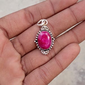 May include: A silver pendant with an oval, faceted, ruby-colored gemstone. The pendant has a decorative border with small, round accents. The bail is a simple loop for a chain. The gemstone is a vibrant, deep red color.