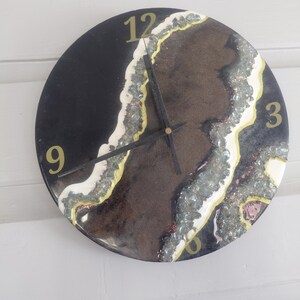 May include: Round black wall clock with gold numerals at 12, 3, 6, and 9. The clock face features a brown and white geode design with silver and yellow accents. The clock hands are black.