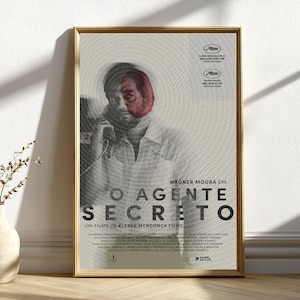 Puede incluir: Un póster de película enmarcado con un marco dorado. El póster presenta el rostro de un hombre con una superposición roja y el título "O AGENTE SECRETO" en letras grandes y en negrita. El póster incluye texto en portugués y logotipos de premios.