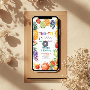 May include: A smartphone displays a colorful birthday invitation with a fruit theme. The text reads "TWO-TTI frutti and such a cutie!" and announces Olivia's 2nd Birthday on Saturday, March 10th at 2:00 PM. The phone rests on a brown card.