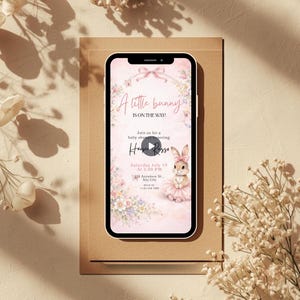 May include: A smartphone displays a baby shower invitation with a pink floral design. The text reads "A little bunny is on the way!" and includes details for the event, featuring a watercolor illustration of a bunny in a tutu.
