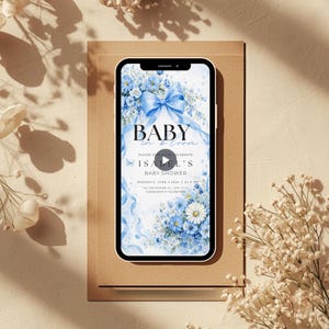 May include: A smartphone shows a baby shower invitation with a blue bow and floral design. The invitation reads "Baby in Bloom" and is for Isabel's baby shower. The event is on June 4, 2023, at 6 PM.