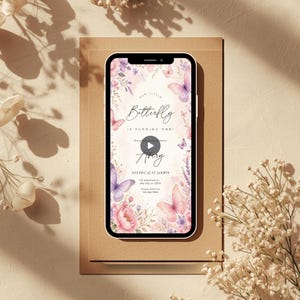 May include: A smartphone displays a digital invitation with a floral and butterfly design. The invitation reads "Our Little Butterfly is Turning One!" with the name "Avery" and event details. The phone rests on a brown surface, with floral accents.