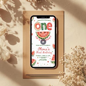 Puede incluir: Un smartphone muestra una invitación de primer cumpleaños con temática de sandía. El diseño presenta la palabra "one" y una rodaja de sandía. La invitación incluye el texto "Olivia's First Birthday" y los detalles del evento. El teléfono está sobre una superficie marrón.
