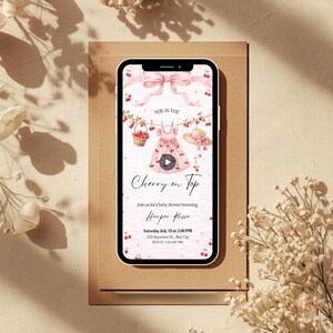 May include: A smartphone displays a baby shower invitation with a cherry theme. The invitation features a pink dress, a straw hat, and the text "Cherry on Top." The design includes cherries, a bow, and the event details.