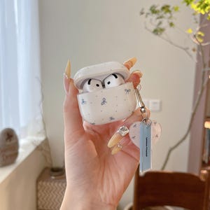 Puede incluir: Funda blanca para AirPods con un patrón de lazo azul, abierta para revelar los auriculares. Un llavero plateado con un dije en forma de corazón y una etiqueta azul que dice "Lemon Crystal" está adjunto.