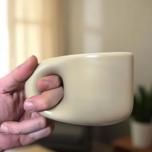 Könnte beinhalten: Eine beige Keramik-Tasse mit rundem Henkel, gehalten von einer Hand. Die Tasse hat eine glatte, matte Oberfläche und ein schlichtes, modernes Design. Die Tasse ist neutralfarben und eignet sich für Kaffee, Tee oder andere Getränke.