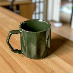 May include: A dark green ceramic mug with a geometric handle. The mug has a cylindrical body and a glossy finish. The mug is sitting on a wooden surface. The brand name "quinnoka" is visible in the top right corner.