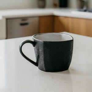 May include: A black ceramic mug with a textured exterior and a white interior sits on a white countertop. The mug has a geometric, faceted design and a curved handle. The kitchen setting includes a sink, cabinets, and plants.