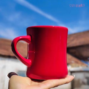 May include: A bright red ceramic mug with a curved handle. The mug is held in a hand against a backdrop of a blue sky and a building. The mug has a glossy finish and a simple, classic design.