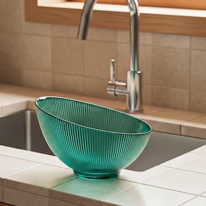 以下が含まれることがあります： キッチンに設置された、ターコイズ色のリブガラスボウル。ボウルは丸みを帯びたベースと角度の付いたリムを備えています。ステンレス製の蛇口と窓が背景にあります。食器や装飾品として使用できます。
