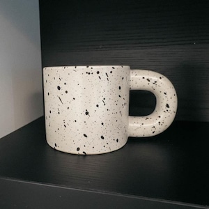May include: A white ceramic mug with black speckles and a rounded handle. The mug is cylindrical and sits on a black surface. The brand name "quinnora" is visible in the upper right corner.