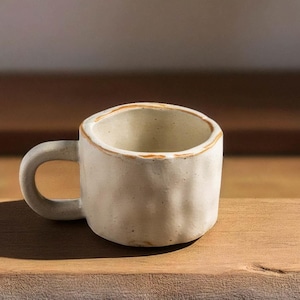 Könnte beinhalten: Eine kleine, cremefarbene Keramiktasse mit rundem Henkel. Die Tasse hat eine strukturierte, leicht unregelmäßige Oberfläche und einen hellbraunen Rand. Sie steht auf einer Holzoberfläche, mit dem Markennamen "quinNora" im Hintergrund.