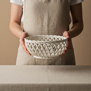 May include: A white, woven ceramic bowl with a lattice design. The bowl is round and held by a person wearing a beige apron. The background is a neutral beige color, and the bowl is sitting on a beige tablecloth.