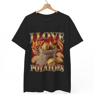 May include: A black short-sleeve t-shirt featuring a graphic print of a burlap sack filled with raw potatoes, surrounded by cooked potato wedges and fries. The text I LOVE POTATOES is printed in large, gold, metallic-style lettering above and below.
