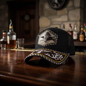Op de afbeelding: Zwarte truckerpet met een bruine en witte decoratieve rand. De pet heeft een zilveren stierembleem op een bruine patch. De pet ligt op een houten bar.