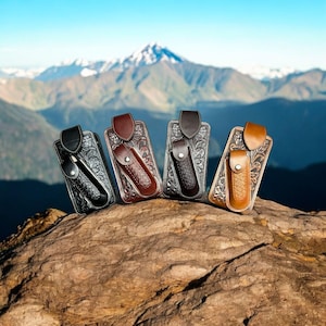 Puede incluir: Cuatro fundas de cuero para cuchillos en negro, marrón y beige, cada una con un patrón floral decorativo. Las fundas se exhiben sobre una roca con un fondo de montaña. Cada funda contiene un cuchillo o herramienta pequeña.