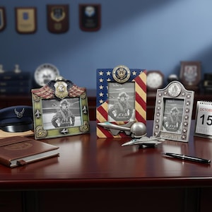 Puede incluir: Tres marcos de fotos decorativos con emblemas militares y diseños patrióticos. Un marco presenta un motivo de bandera estadounidense, otro tiene un diseño de águila y alas, y el tercero tiene un patrón de estrellas. También hay un modelo de avión en miniatura y un bolígrafo sobre la mesa.