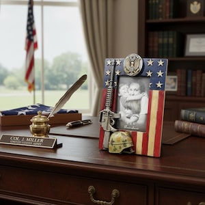 Könnte beinhalten: Ein patriotischer Fotorahmen mit amerikanischem Flaggenmuster, einer Miniaturwaffe und einem Militärhelm. Der Rahmen fasst ein 10x15 cm großes Foto. Ein Tintenfass, ein Stift und ein Namensschild befinden sich auf dem Schreibtisch. Im Hintergrund ist eine amerikanische Flagge zu sehen.