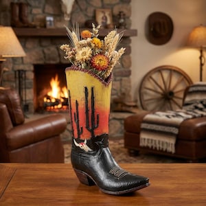 May include: A decorative cowboy boot vase with a desert sunset design. The boot is black with a yellow and red gradient upper, featuring cacti and a skull. It holds dried flowers and wheat stalks, adding a rustic touch to home decor.