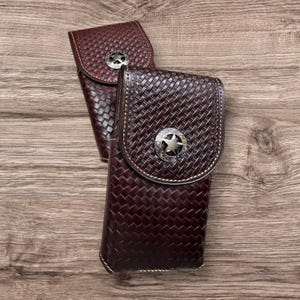May include: Two burgundy leather phone cases with a woven texture and silver star-shaped embellishments. The cases have a flap closure and are stitched with a light-colored thread. The background is a wooden surface.