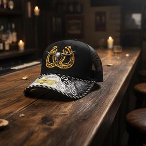May include: Black trucker hat with a mesh back and a decorative brim. The hat features a gold horseshoe and bull skull design on the front. The brim has a black and white pattern.