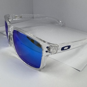 May include: Clear-framed sunglasses with vibrant blue lenses. The sunglasses have a rectangular shape and a transparent frame. The lenses reflect a bright blue color. The temples have a small logo. The sunglasses are designed for eye protection.