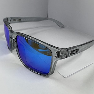 May include: Gray sunglasses with blue lenses. The frames are a translucent gray, and the lenses reflect a bright blue hue. The sunglasses have a rectangular shape and a modern design, suitable for outdoor activities.