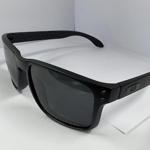 May include: Black sunglasses with a square frame and dark lenses. The sunglasses have a matte finish and a subtle logo on the temples. The arms curve slightly for a comfortable fit. These sunglasses are suitable for outdoor activities.