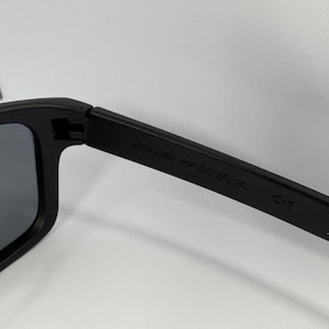 May include: Close-up of black sunglasses with dark gray lenses. The arm of the sunglasses has the text "MADE IN U.S.A." and "CE" printed on it. The sunglasses are set against a white background.
