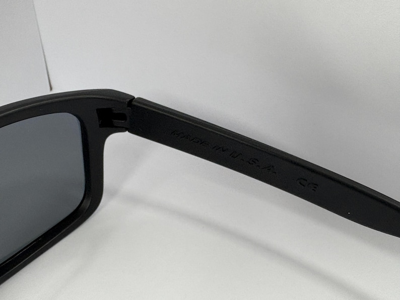 May include: Close-up of black sunglasses with dark gray lenses. The arm of the sunglasses has the text "MADE IN U.S.A." and "CE" printed on it. The sunglasses are set against a white background.