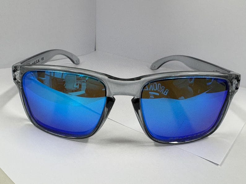 May include: Gray sunglasses with blue mirrored lenses. The sunglasses have a square frame and a clear gray plastic frame. The lenses reflect the surrounding environment. The sunglasses are on a white surface.