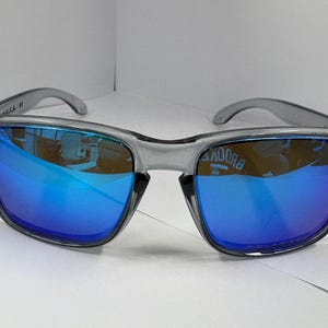 May include: Gray sunglasses with blue mirrored lenses. The sunglasses have a square frame and a clear gray plastic frame. The lenses reflect the surrounding environment. The sunglasses are on a white surface.