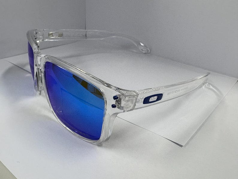 May include: Clear-framed sunglasses with vibrant blue lenses. The sunglasses have a rectangular shape and a transparent frame. The lenses reflect a bright blue color. The temples have a small logo. The sunglasses are designed for eye protection.