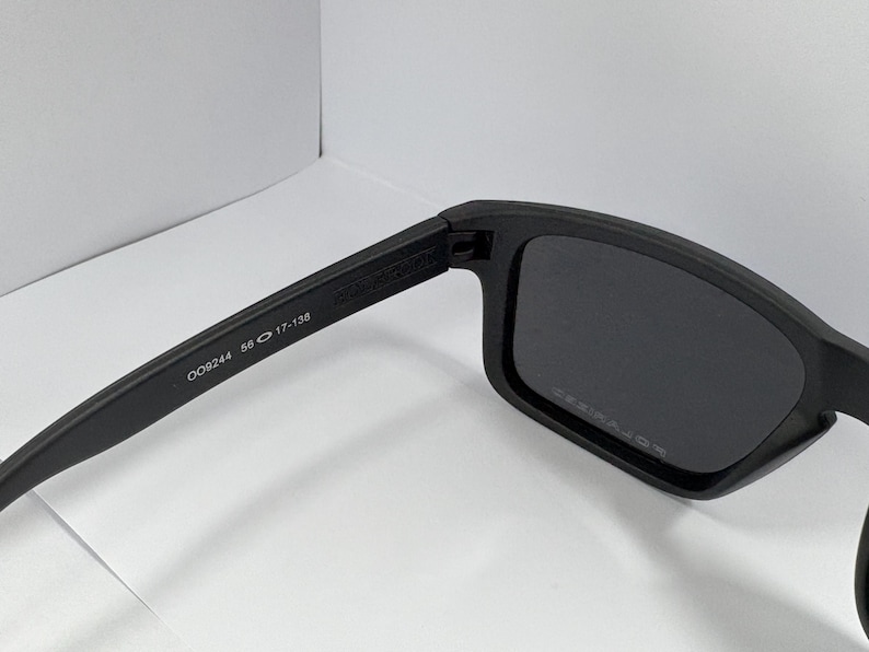 May include: Black sunglasses with rectangular frames. The left arm of the sunglasses has the numbers "009244 56 17-138" printed on it. The lenses are dark, and the frames are matte black.
