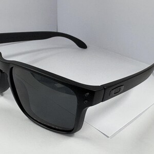May include: Black sunglasses with a square frame and dark lenses. The sunglasses have a matte finish and a subtle logo on the temples. The arms curve slightly for a comfortable fit. These sunglasses are suitable for outdoor activities.