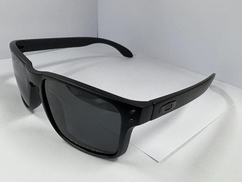 May include: Black sunglasses with a square frame and dark lenses. The sunglasses have a matte finish and a subtle logo on the temples. The arms curve slightly for a comfortable fit. These sunglasses are suitable for outdoor activities.